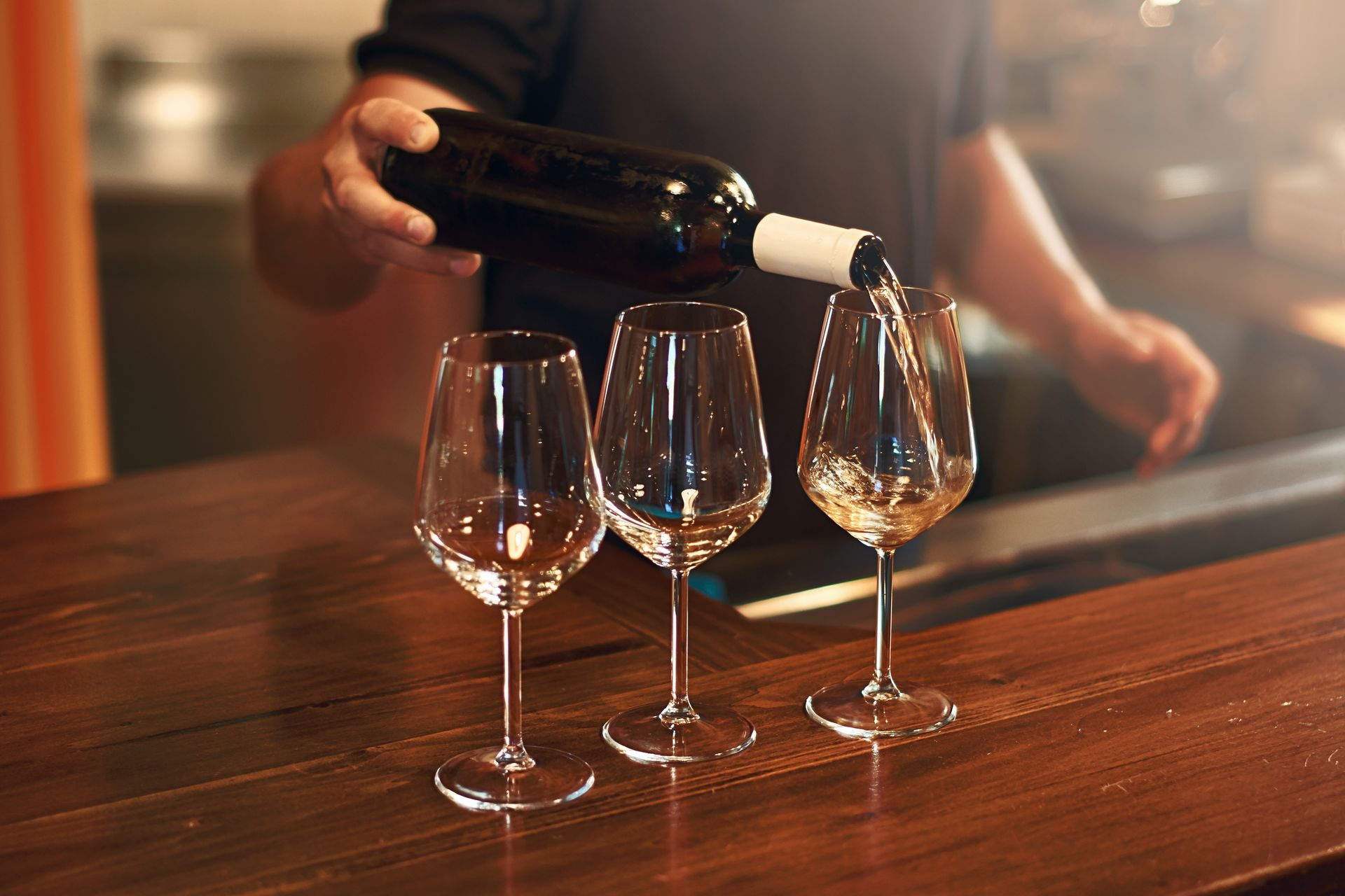 A man is pouring wine into three glasses on a wooden table.