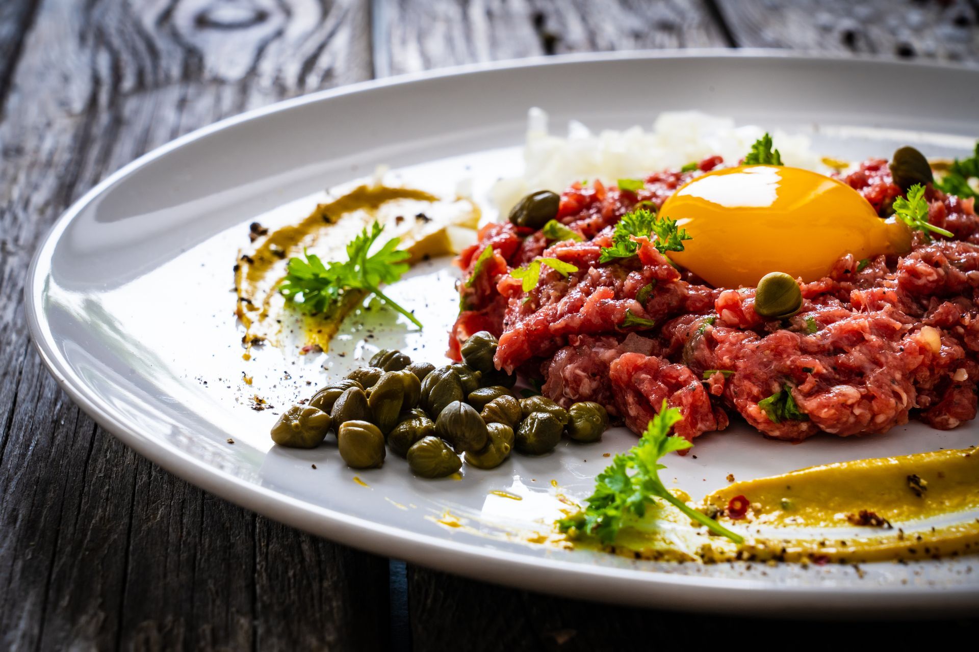 A plate of food with meat , eggs , capers and mustard on a wooden table.