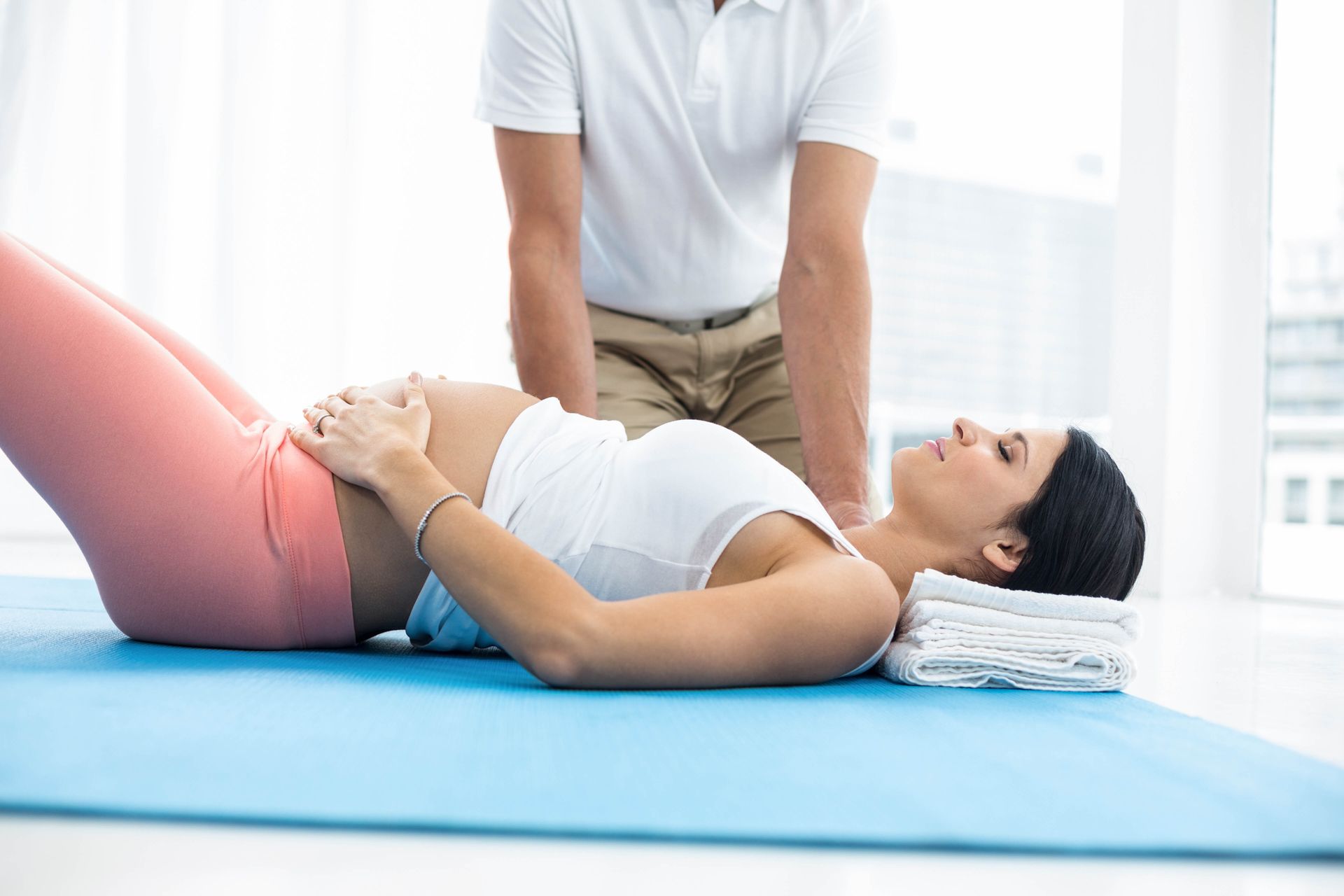 Pregnant woman lying down with a therapist, hands on belly, white top, pink pants, blue mat.