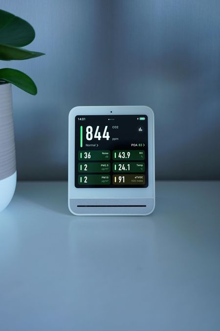 Air quality monitor displays readings; white device on a table with a plant in the background.