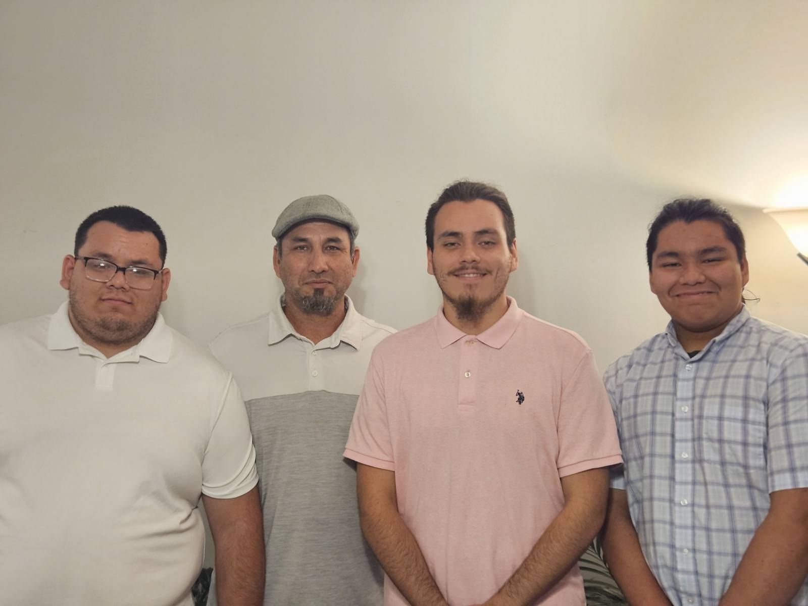 Four men stand in front of a white wall. They are wearing casual shirts, with one wearing a hat. All are smiling.