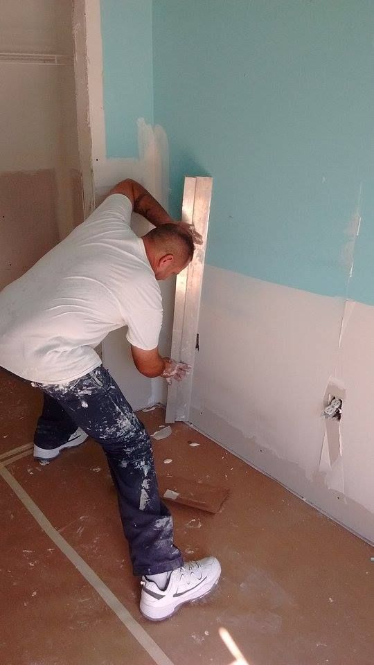 Man in paint-splattered jeans and white shirt installing wooden trim in a room with blue and white painted walls.