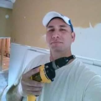 Man in a white t-shirt and cap, holding a yellow and black power drill; he appears to be working indoors.