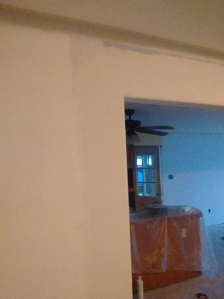 White wall with doorway leading to a room with a ceiling fan and kitchen cabinets covered in plastic.