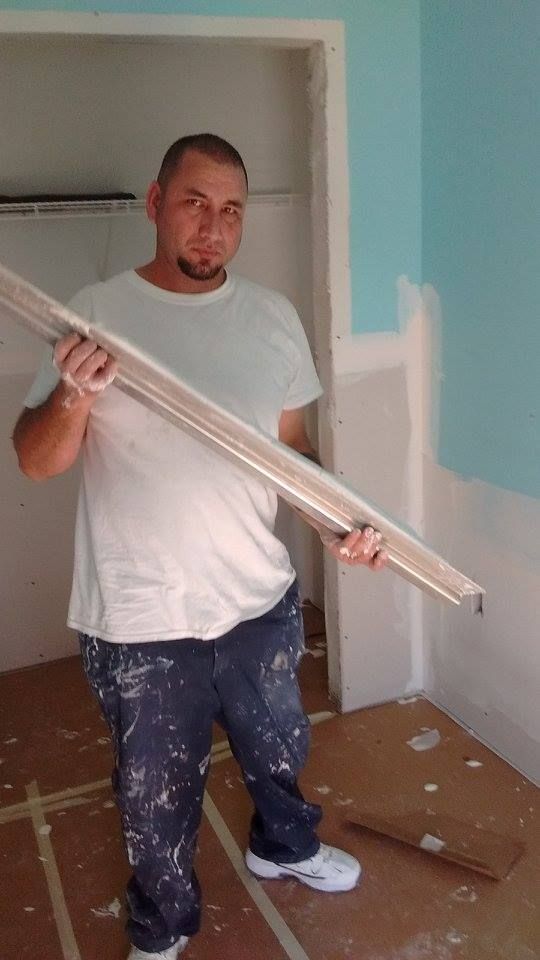 Man holding wood trim in a room under construction, wearing paint-splattered jeans and a white t-shirt.