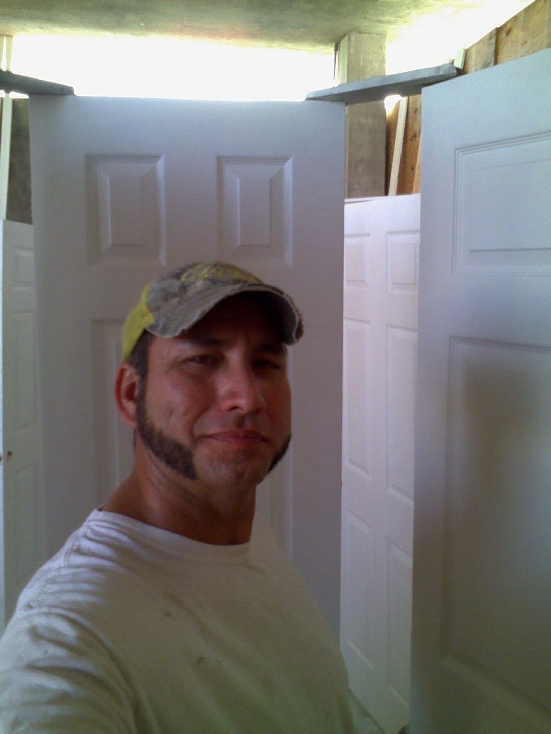 Man with a camo hat and goatee stands in front of several white panel doors. He wears a white t-shirt, and smiles slightly.