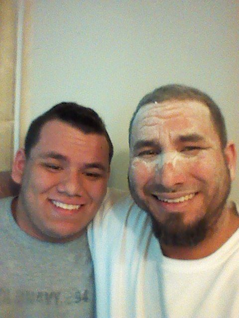 Two men smiling, embracing. One with white powder on his face, the other with a grey shirt. Inside.