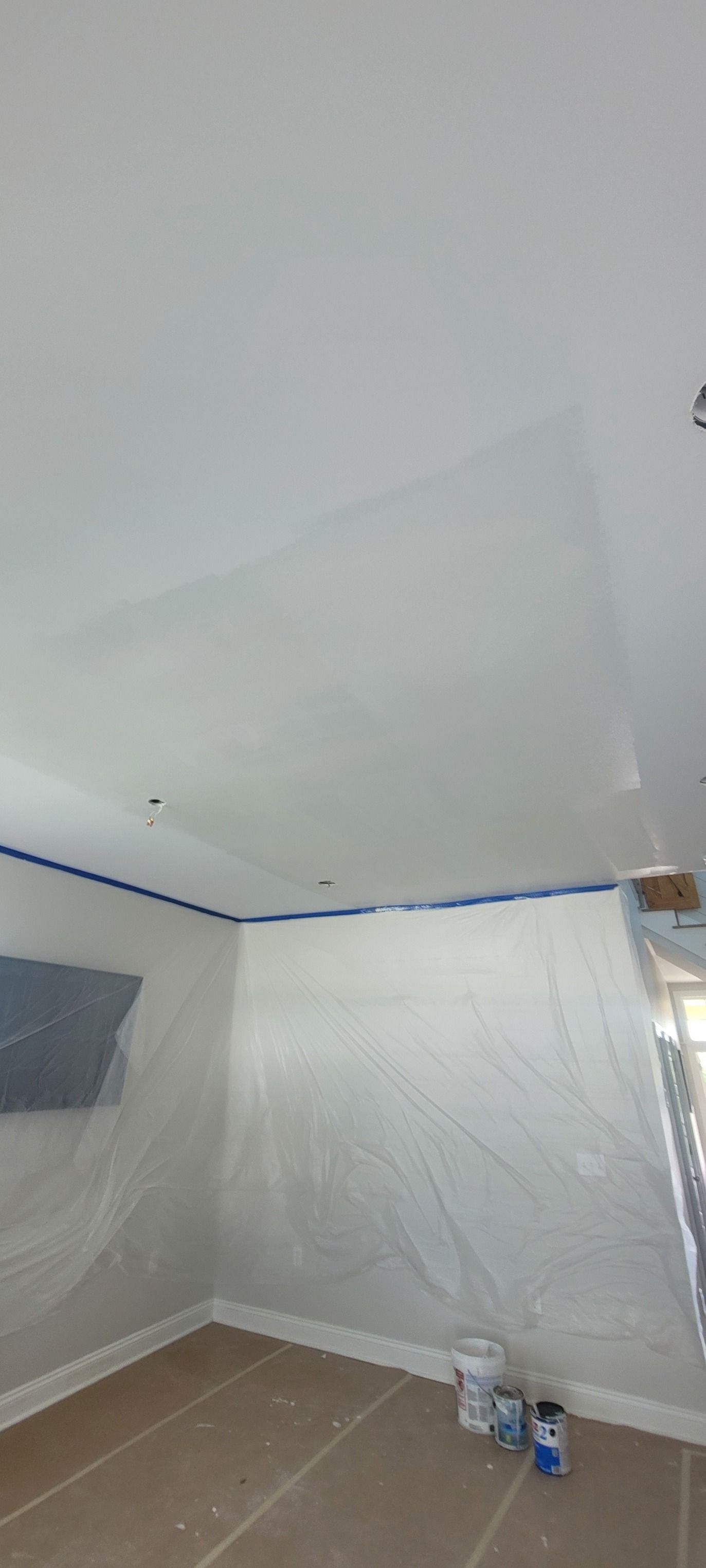 Room prepared for painting, walls and floor covered in plastic sheeting. Blue tape lines the ceiling edges.