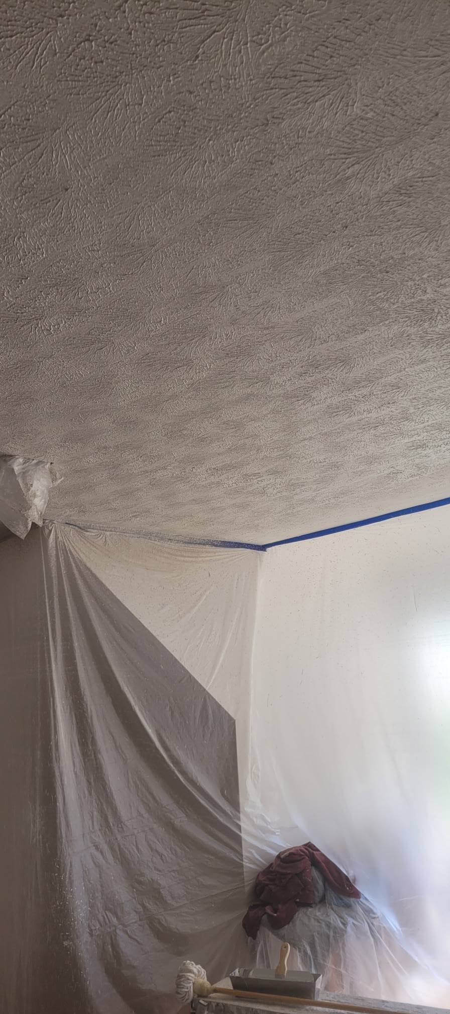 A room with plastic sheeting protecting walls during ceiling work. Blue painter's tape lines the ceiling edges.