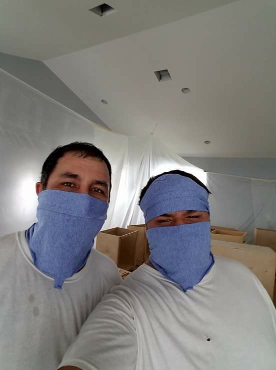 Two men in white shirts and blue face coverings posing in a room with white walls and cardboard boxes.