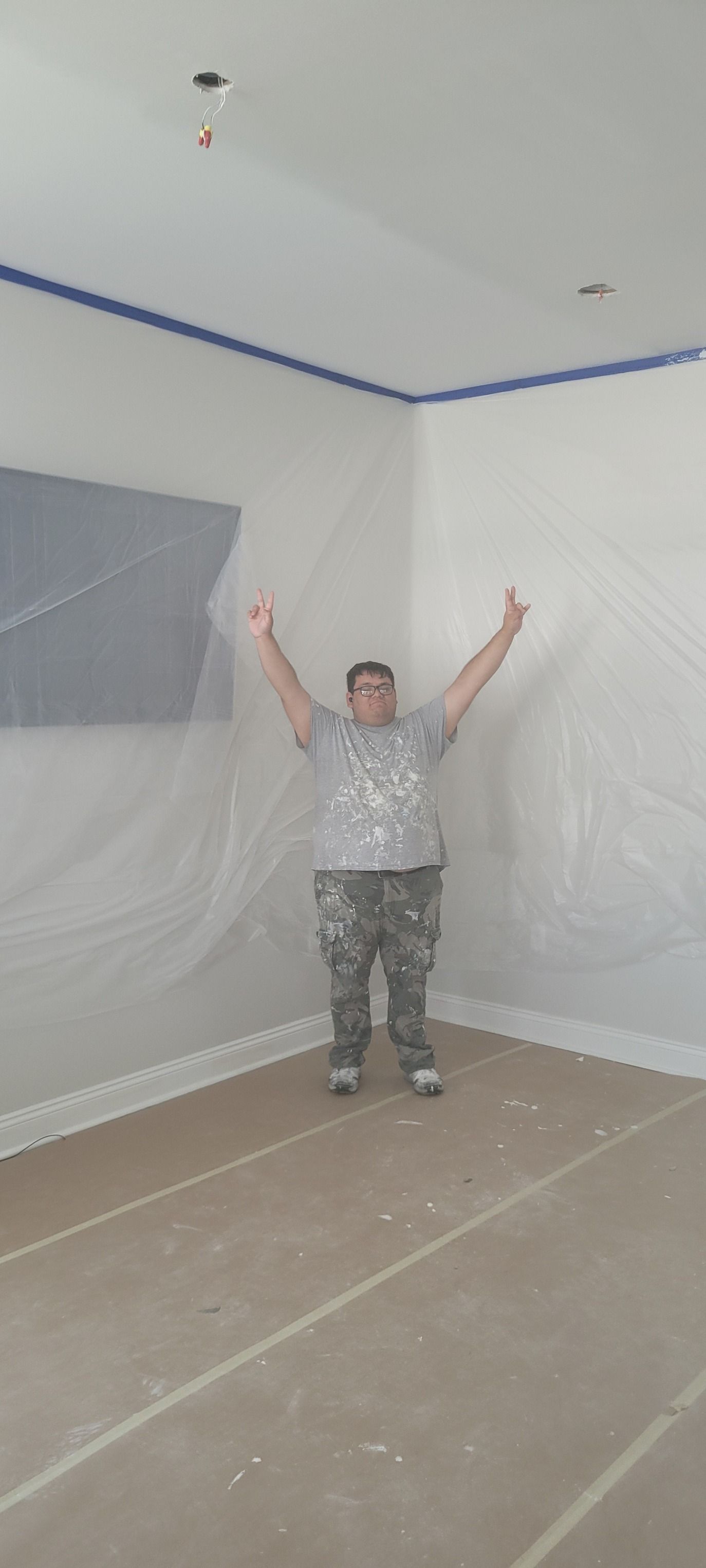 Man standing in a room, arms raised, ready to paint. Walls are taped off; the floor is covered.