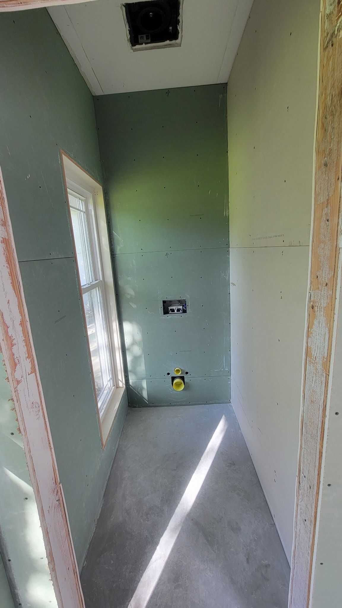 A narrow, unfinished room with green drywall, a window on the left, and a light fixture and pipe visible.
