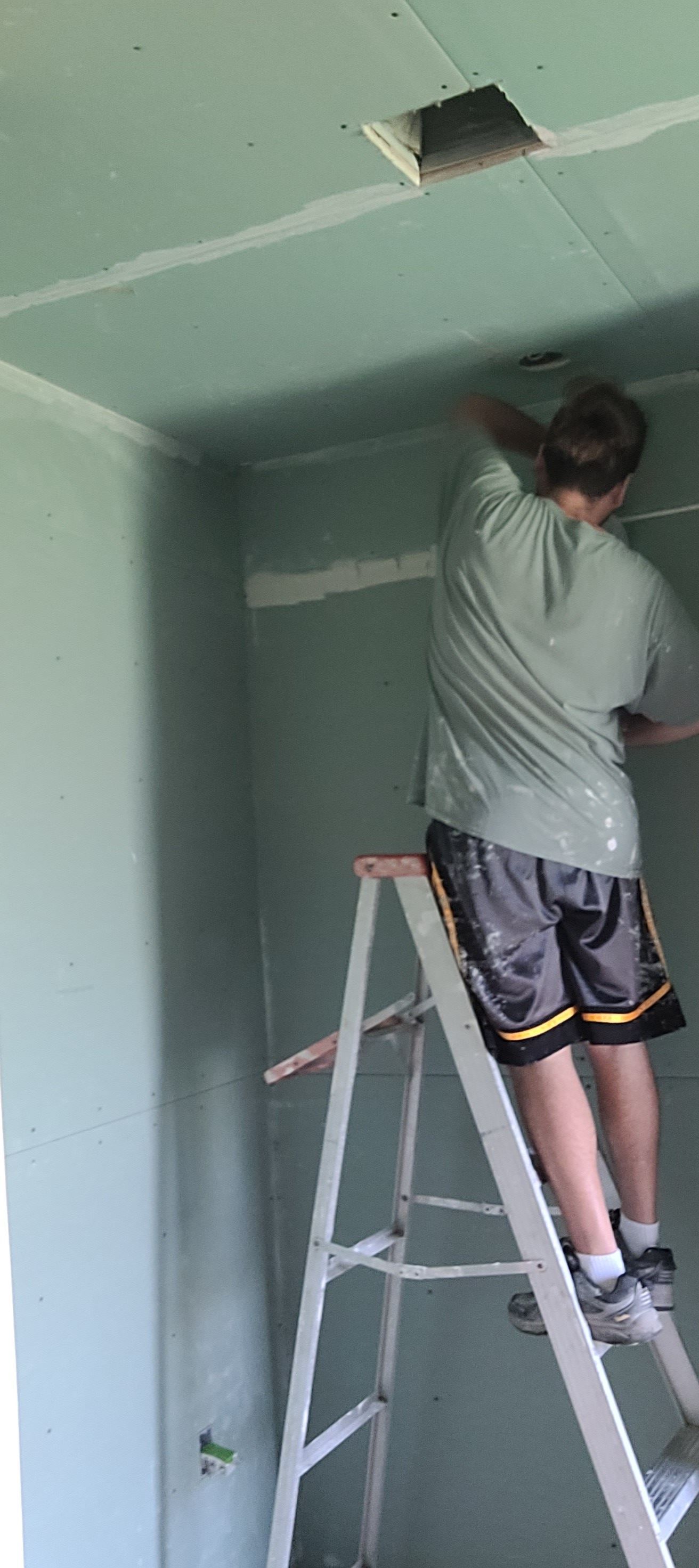 A person on a ladder works on the ceiling of a room that's being renovated. The walls and ceiling are a pale green.