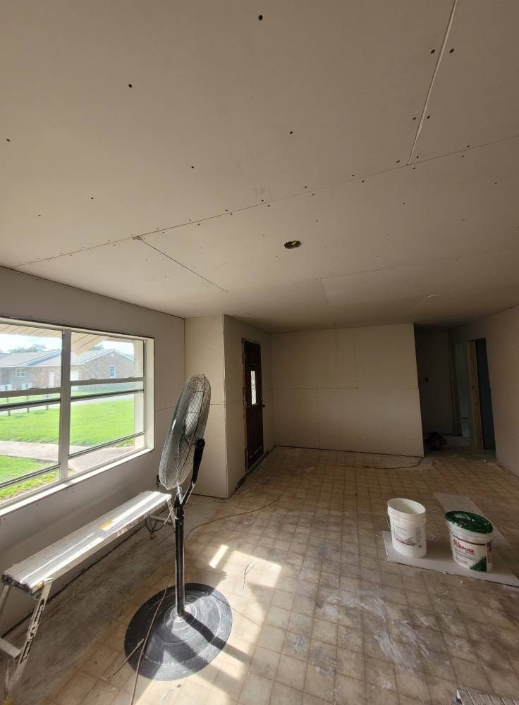 An empty room under construction. The room has drywall walls and ceiling, a window, and a standing fan.