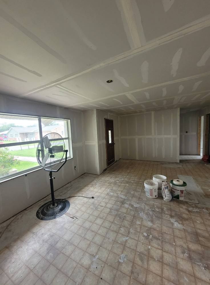Interior room with drywall in progress, a fan, buckets, and a window with an outside view. The floor is tiled and worn.