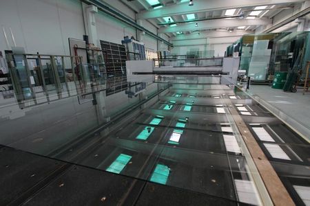 Grandi lastre di vetro appoggiate su un tavolo da taglio industriale all'interno di uno stabilimento di produzione ben illuminato.