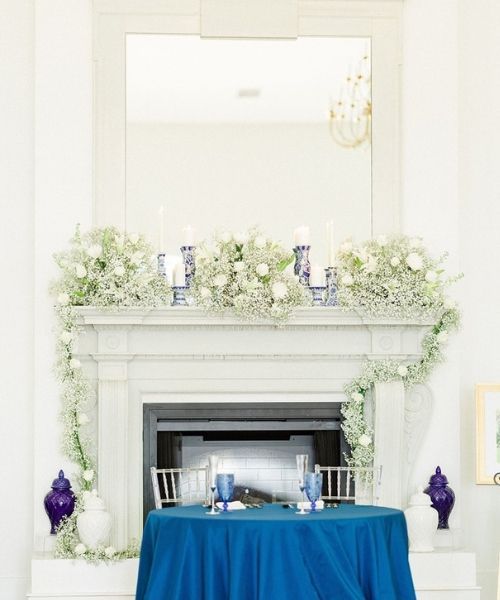 White mantel decorated with green-and-white flowers, candles, and blue accents, with a blue-draped table in front
