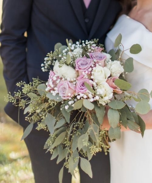 Wedding bouquet of pink and white roses with greenery held by a couple in formal attire