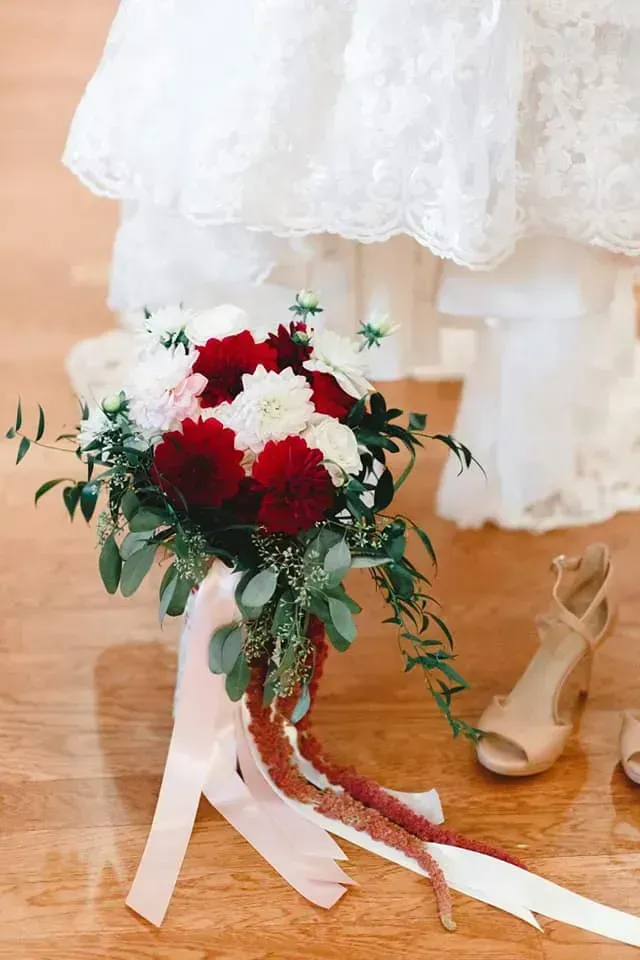 Wedding bouquet with red and white flowers and long ribbons beside a white dress and beige heels on a wooden floor