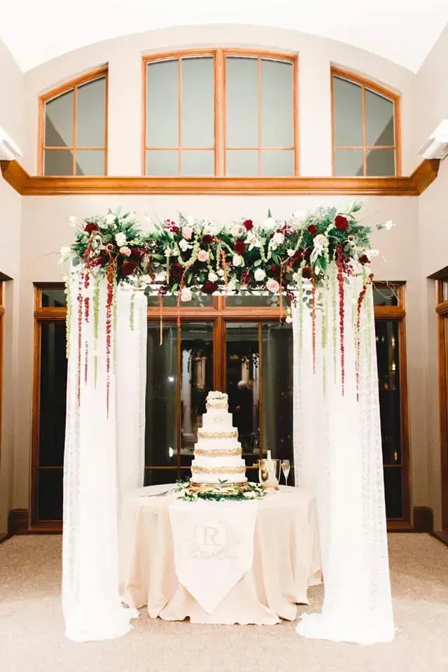 Wedding reception table with white drapes, floral arch, and tiered cake in a bright hall