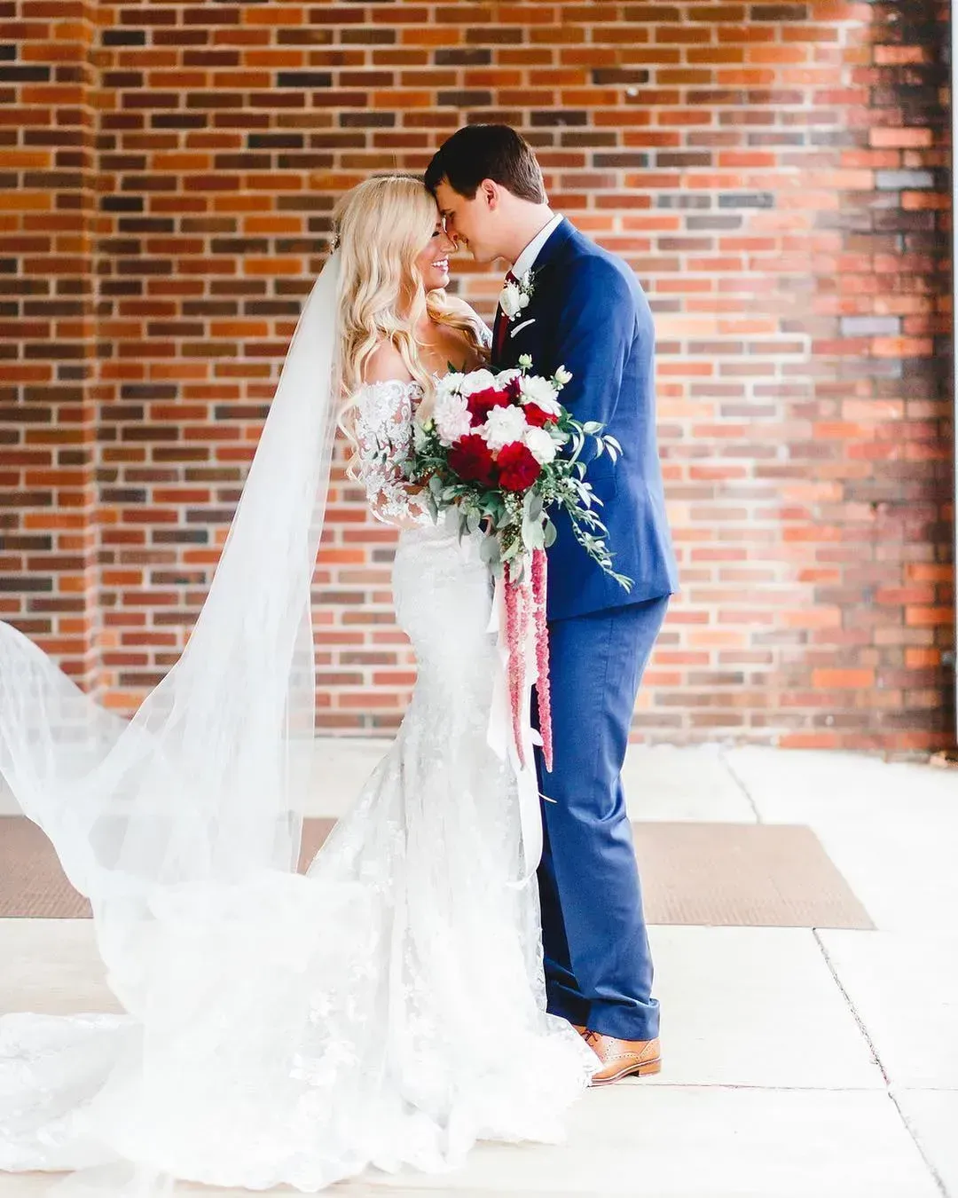 Bride and groom kiss beside a brick wall, holding a red-and-white bouquet.