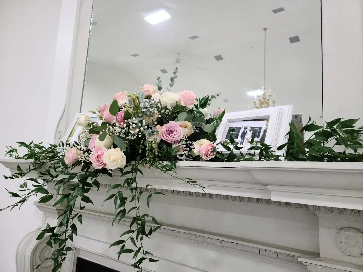 White mantel decorated with pink and cream flowers, trailing greenery, and a framed photo in front of a mirror