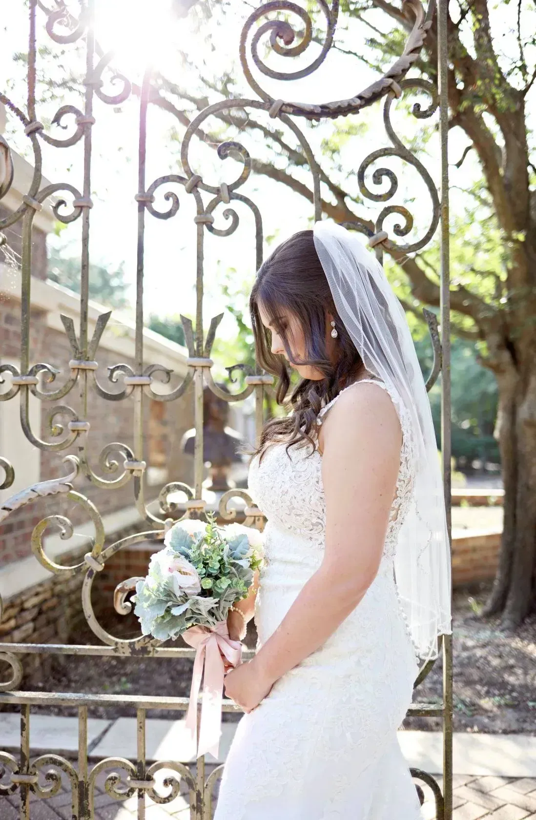 Bride in white dress and veil holding bouquet beside ornate wrought-iron gate outdoors