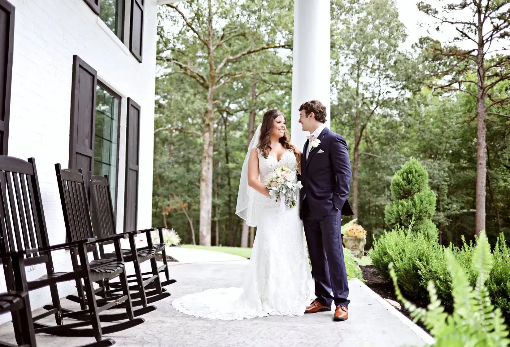 Bride and groom standing on a stone path outside a white house among tall pine trees