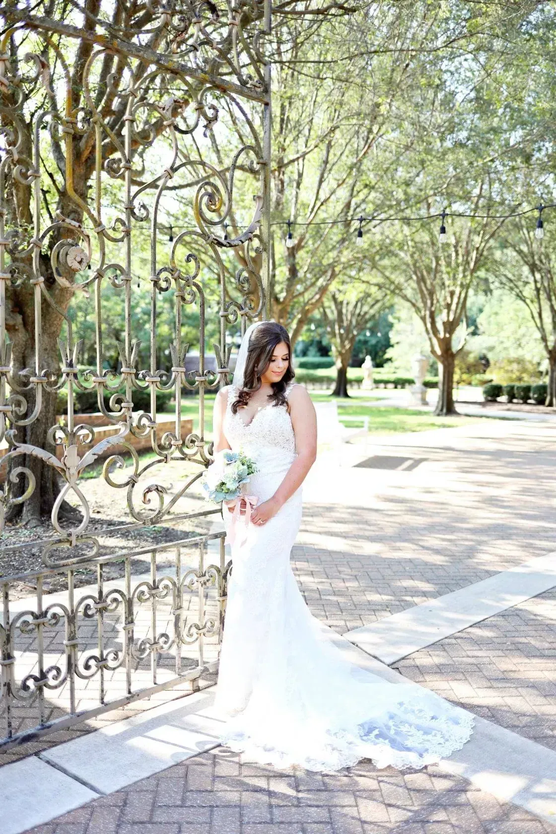 Bride in a white lace gown holding a bouquet beside an ornate gate in a sunlit garden path.
