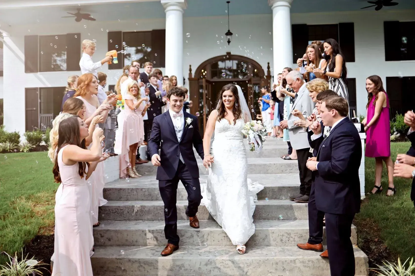 Wedding couple walking down steps as guests cheer outside a white-columned venue