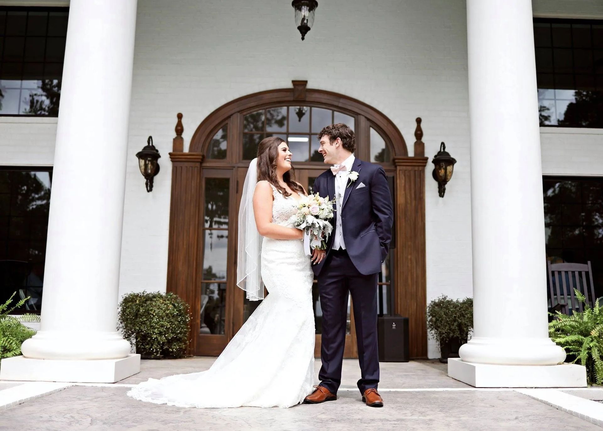 Bride and groom posing outside a white-columned building, holding hands and smiling