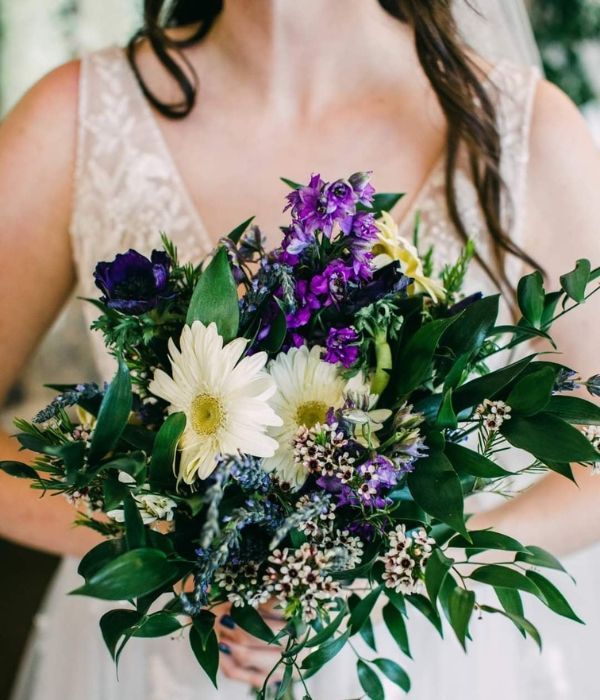 Bride holding a bouquet of white, purple, and green flowers against a lace wedding dress