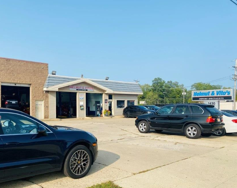 Sunny parking lot outside a small storefront with parked black SUVs and a blue business sign.