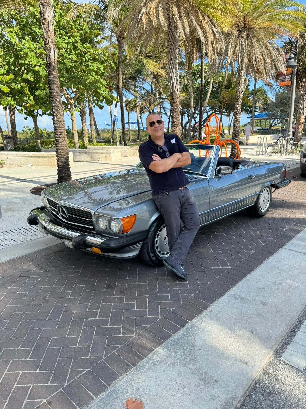 Man with arms crossed leans against a silver convertible car, on a bricked street with palm trees.