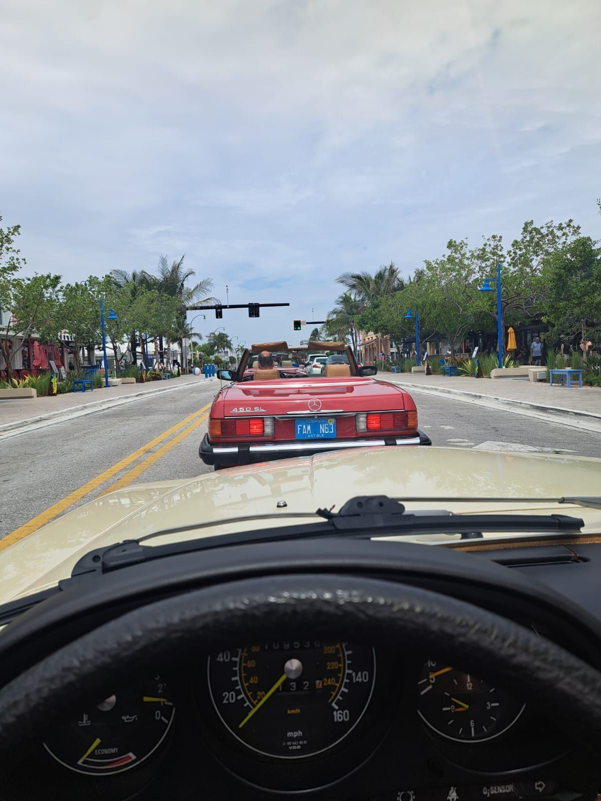 View from car dashboard, following a red convertible on a sunny street lined with trees and shops.