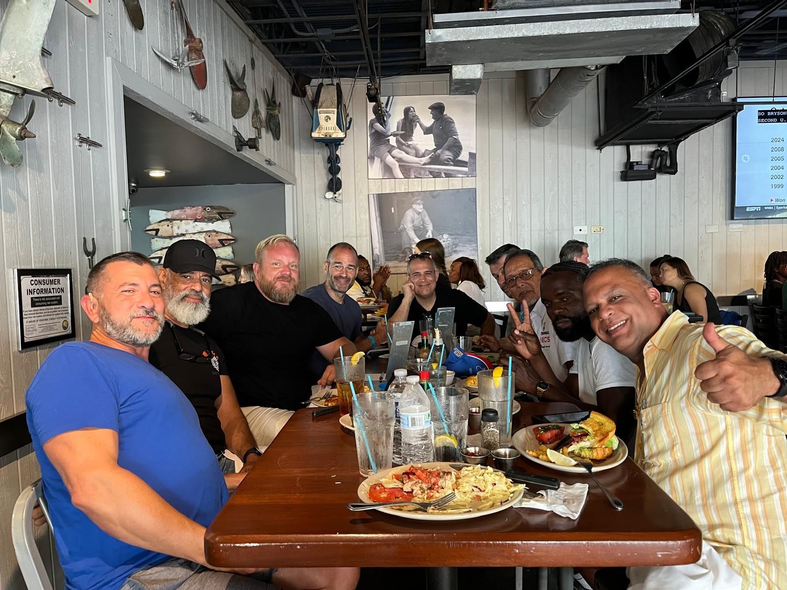 Group of people at a restaurant, seated around a table with food and drinks. Some smile, giving thumbs up.
