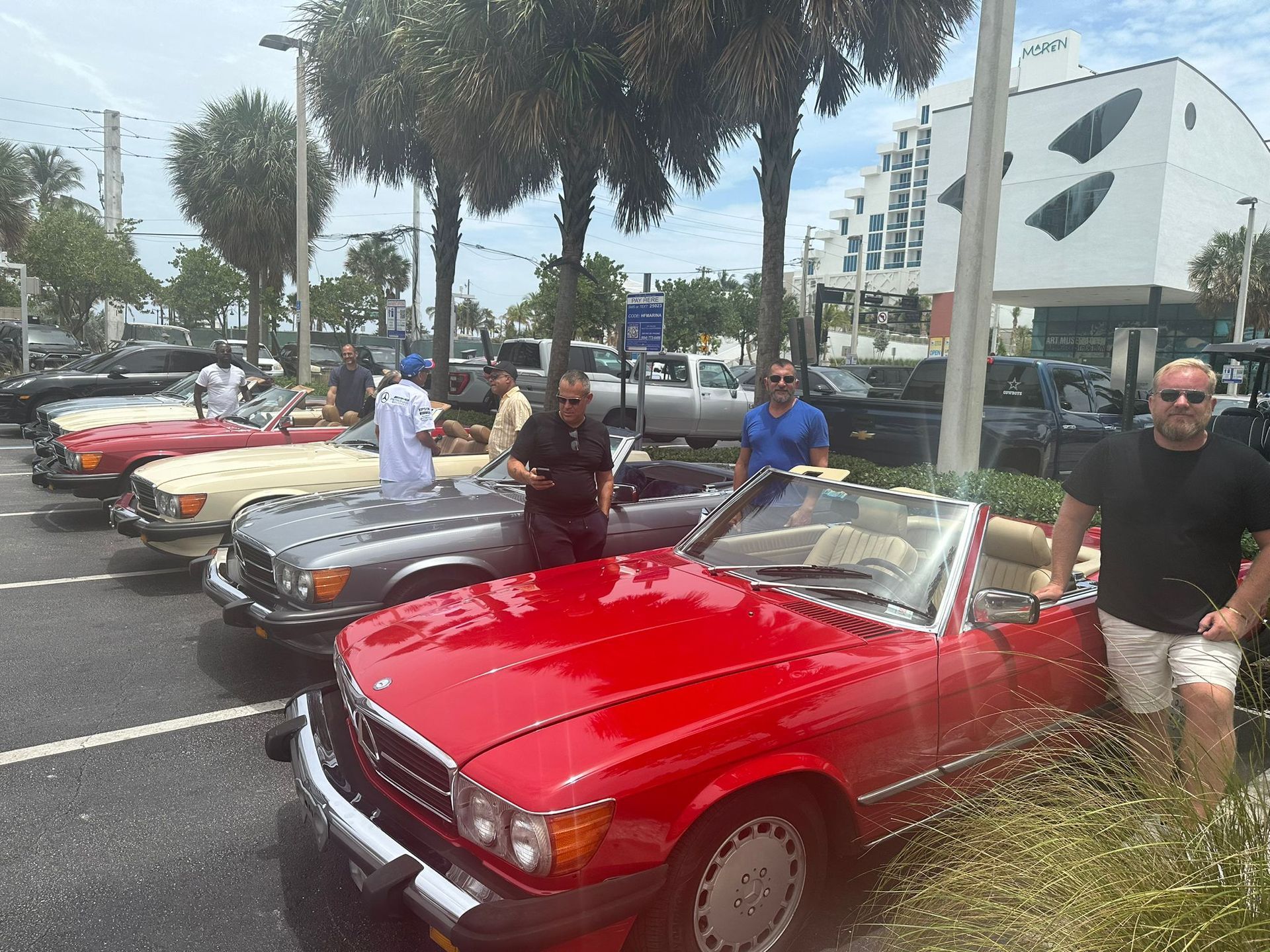 Classic red Mercedes convertible with a group of people at an outdoor gathering, other classic cars in background.