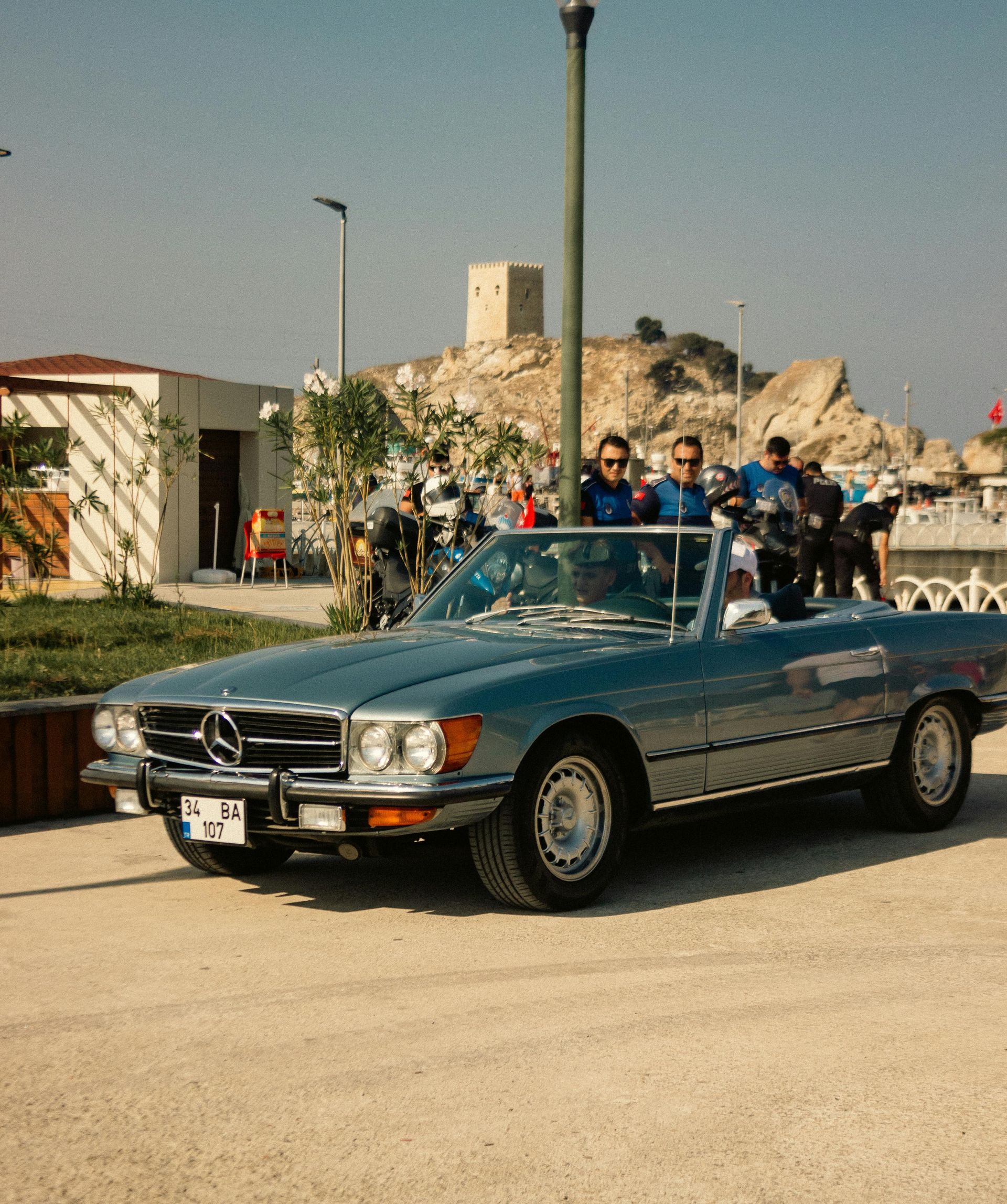 Blue Mercedes-Benz convertible with two men driving past a coastal town with a castle on a hill.