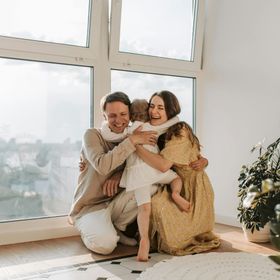 Family hugging by a bright window: smiling parents embrace a child in a sunny room.