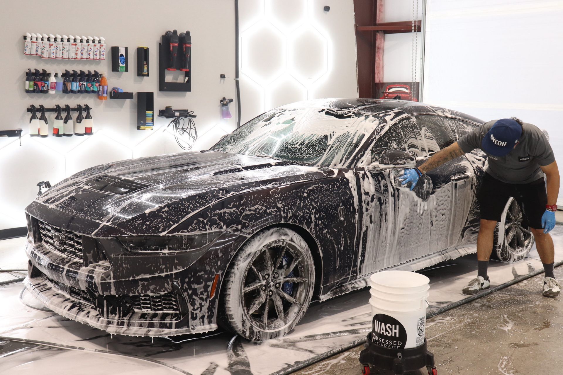 Black car being washed in a garage, covered in foam while a person scrubs the side.