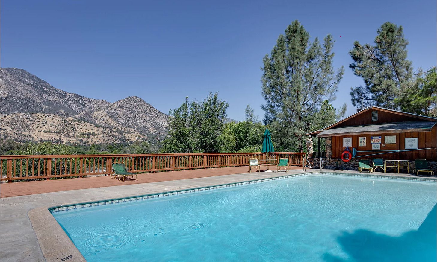 Swimming pool with mountain view, surrounded by a wooden deck and trees, under a blue sky.