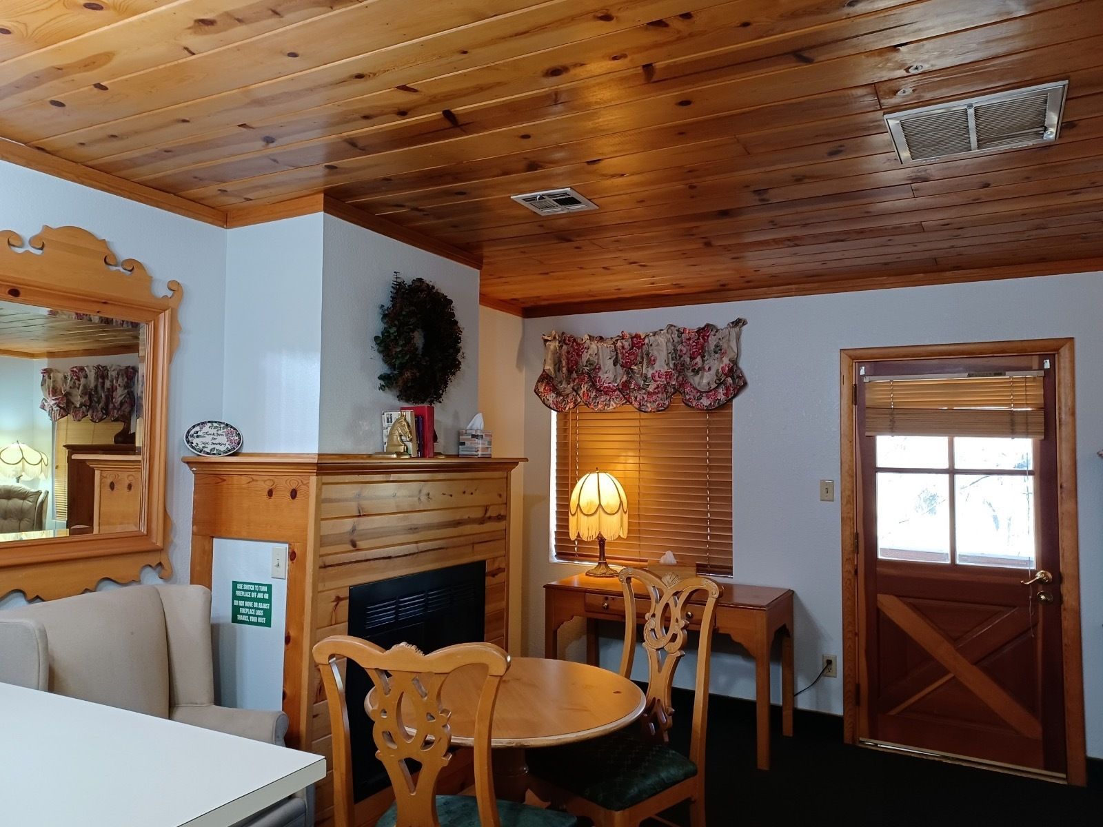 Cozy cabin interior: wood ceiling, fireplace, round table with chairs, window with blinds and valance, door.