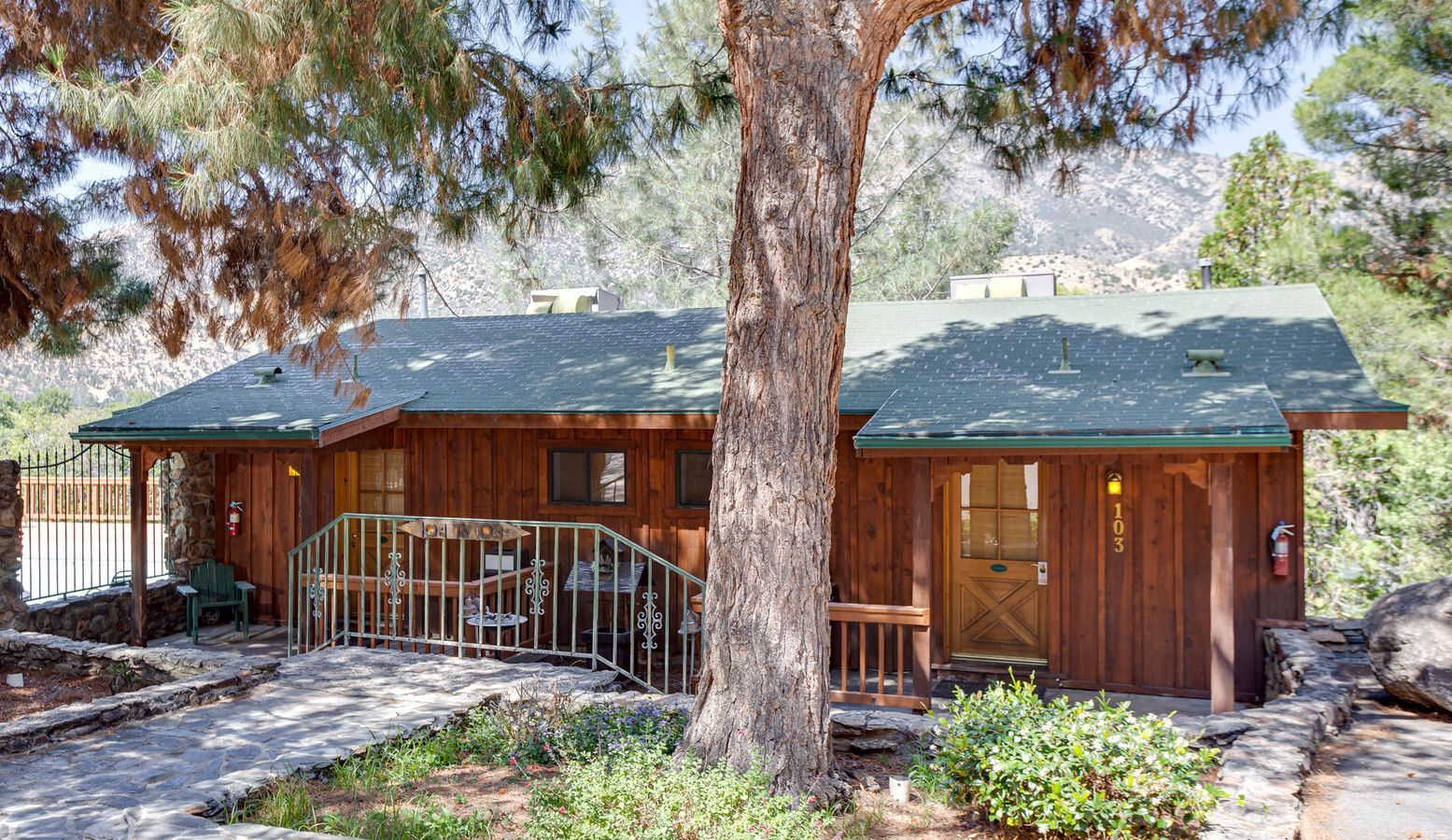 Brown cabin with porch, path, and tree in front, with mountains in the background.