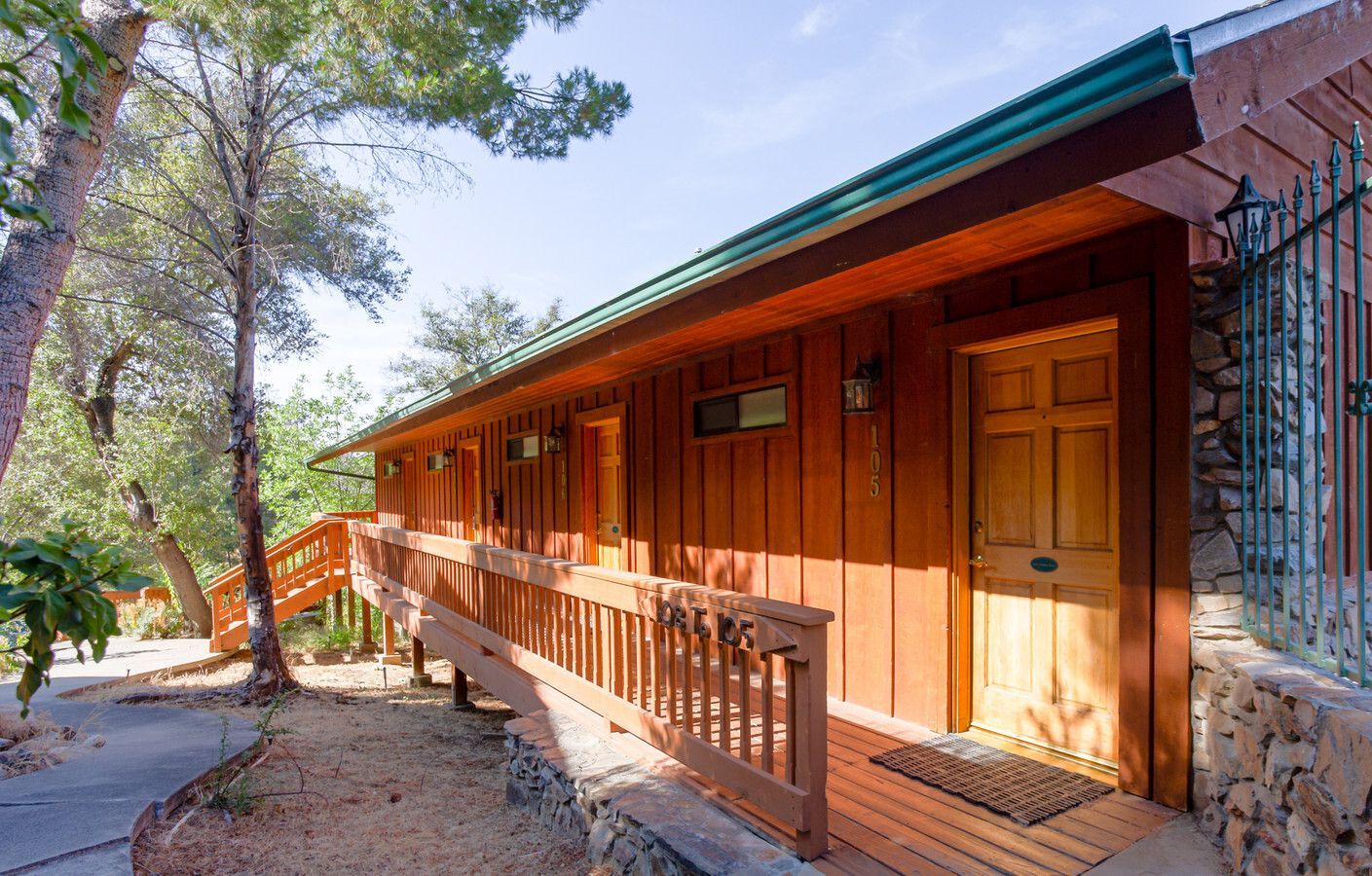 Wooden cabin with a ramp and railing, door, green roof, and stone wall.