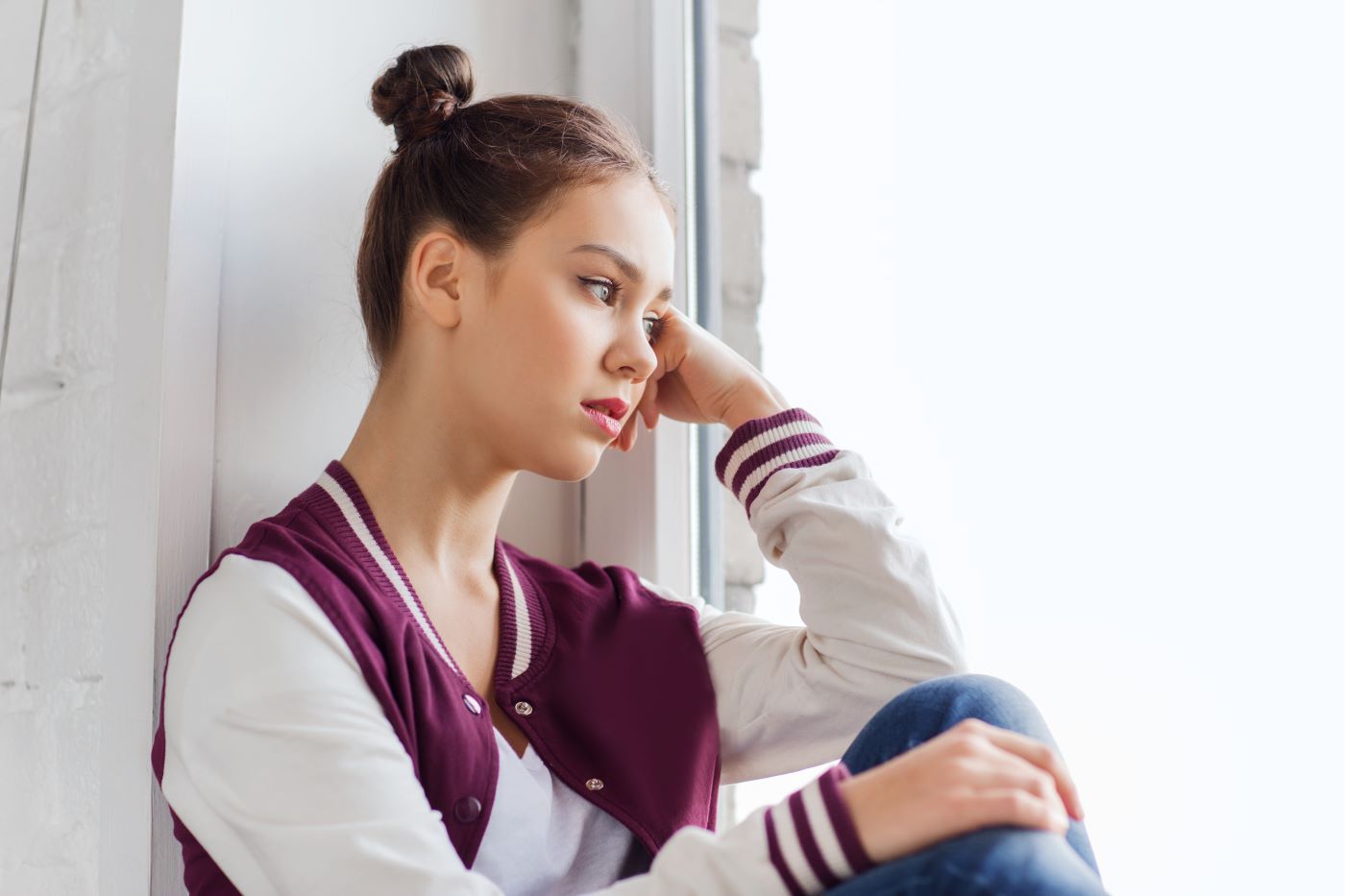 Woman with bun, sitting in a window, looking pensive, wearing a letterman jacket and jeans.