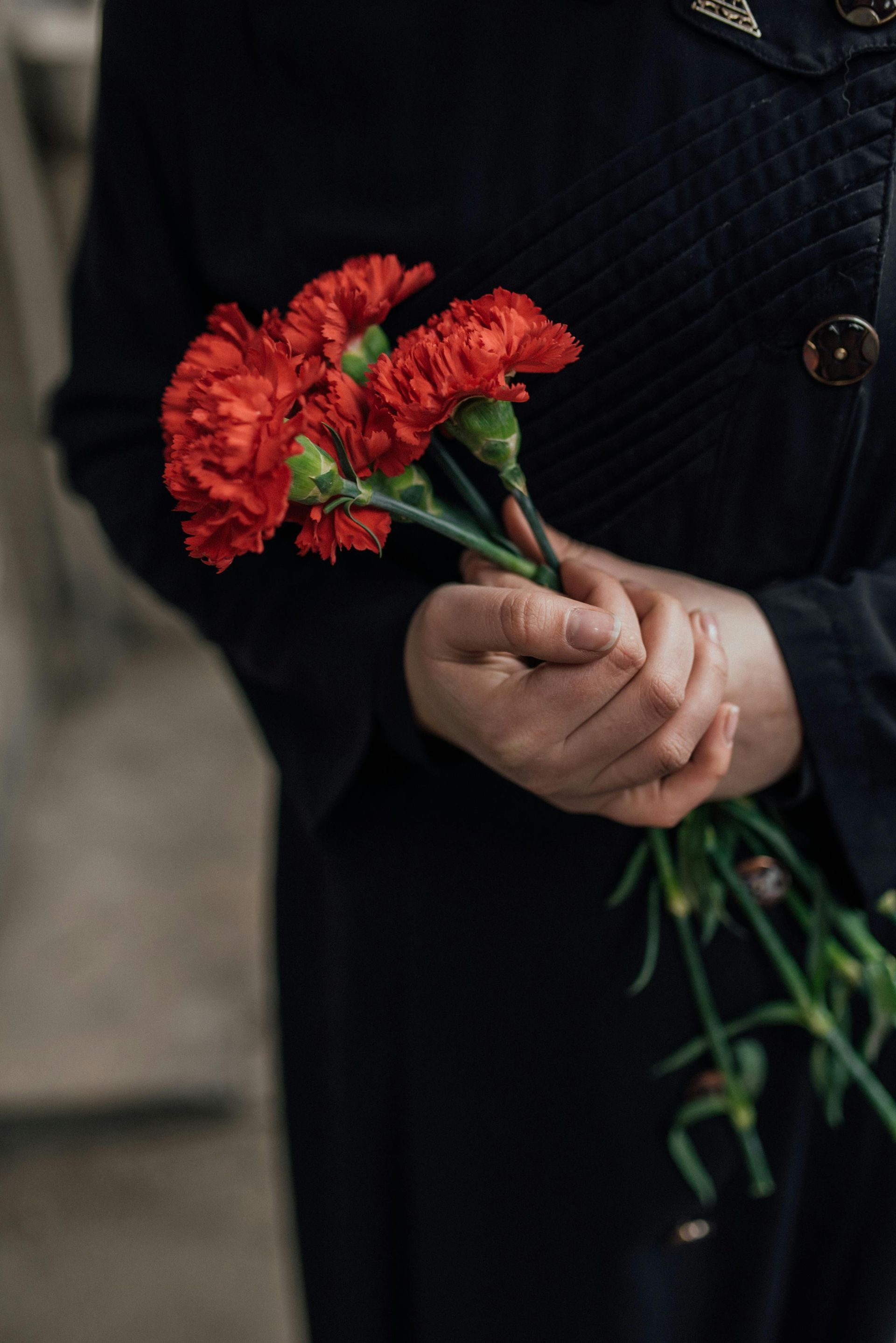 Person in black coat holds red carnation flowers.