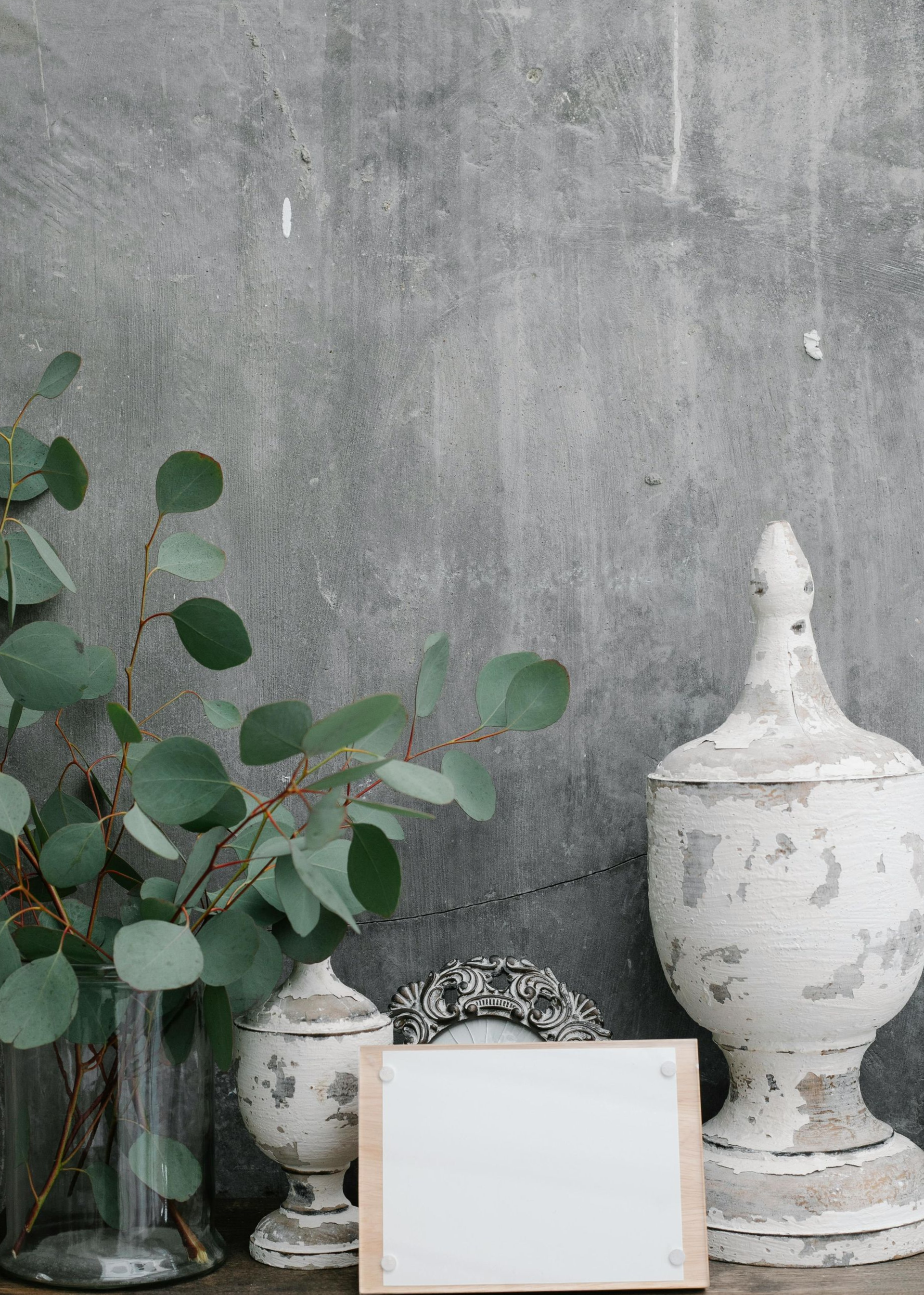 Eucalyptus branches in glass vase, aged white urns, blank card, against a textured gray wall.