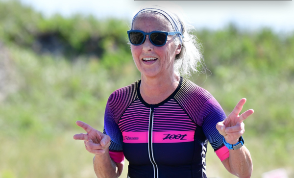 Woman in athletic wear smiles, giving peace signs. Outdoors, near greenery.