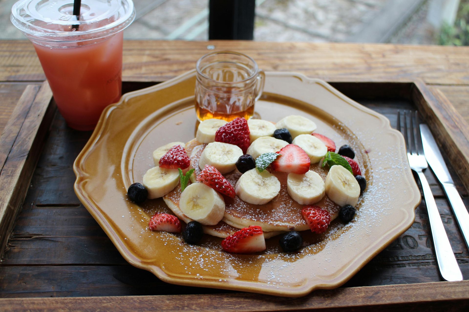 Pancakes topped with berries and bananas, with syrup, juice, and silverware on a wooden tray.