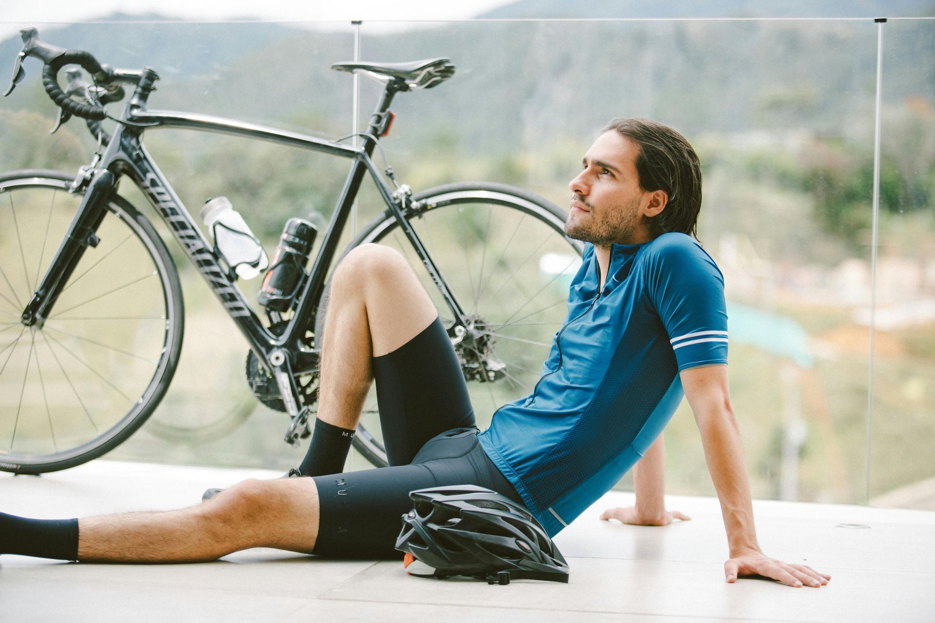 Cyclist resting on patio, bike behind him. Blue jersey, black shorts, helmet. Outdoors with scenic view.